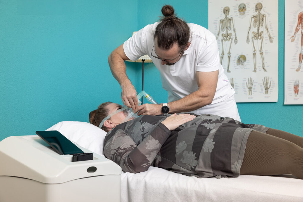 Therapeut bei der Anziehen einer Atemmaske für die IHHT Zelltherapie an eine liegende Patientin in einem Behandlungsraum mit türkisen Wänden und Anatomie-Postern.